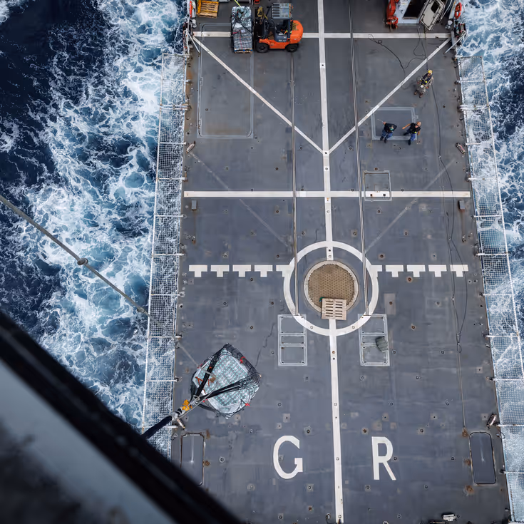 Het grijze helikdek gezien vanuit de helikopter, terwijl het schip vaart.