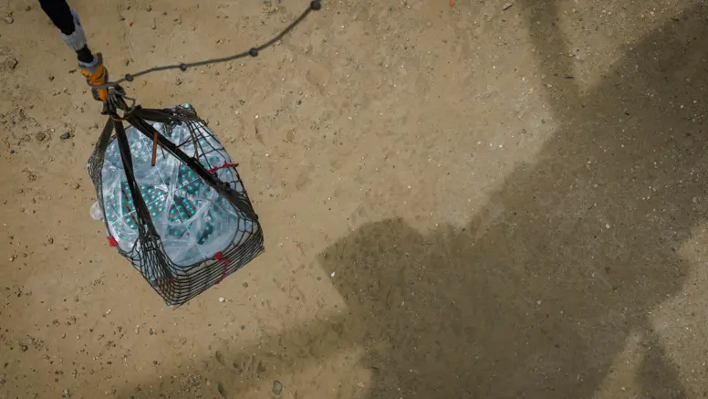 Een pallet waterflessen hangt boven een zandgebied. De kabel zit vast aan een helikopter, waarvan alleen de schaduw is te zien.