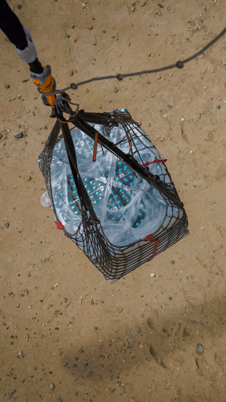 Een pallet waterflessen hangt boven een zandgebied. De kabel zit vast aan een helikopter, waarvan alleen de schaduw is te zien.