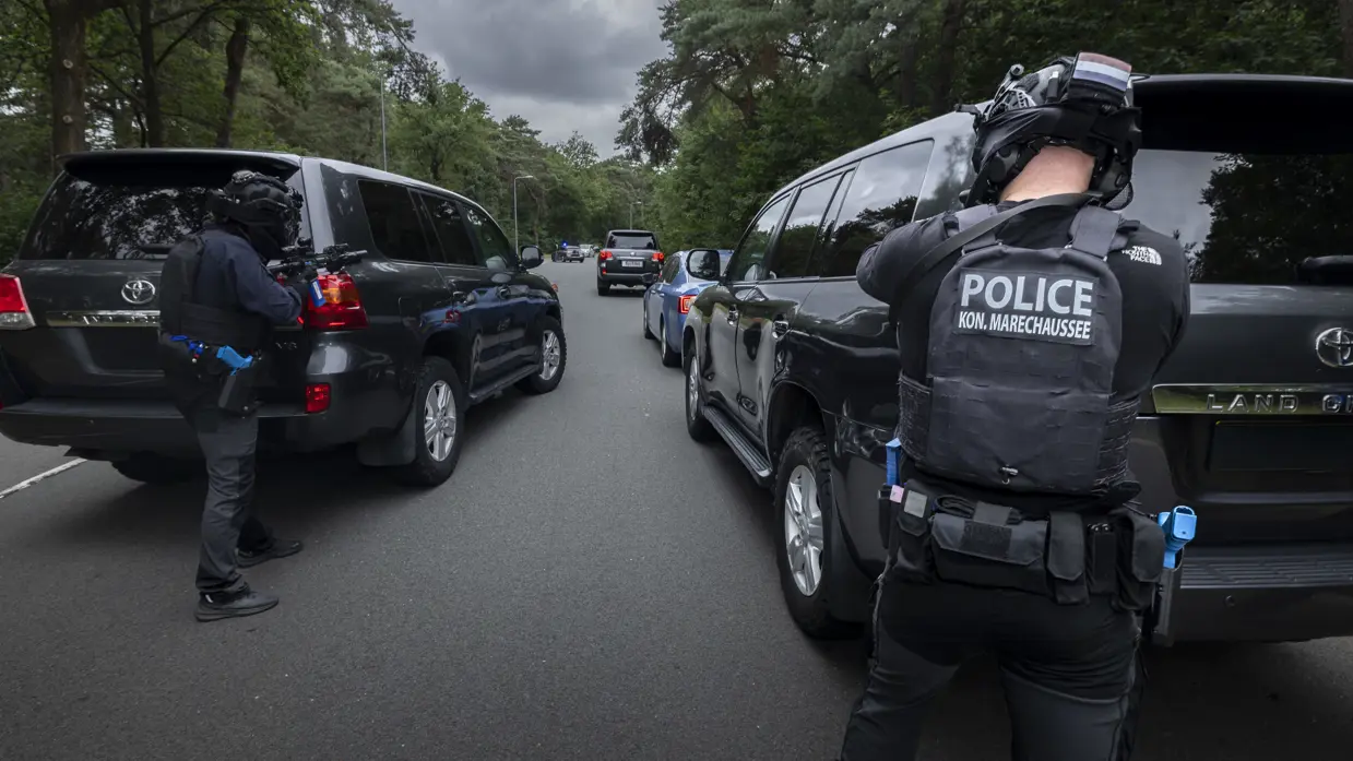 Twee zwaarbewapende cursisten richten elk vanachter een auto met hun vuurwapen op een ingesloten blauwe auto.