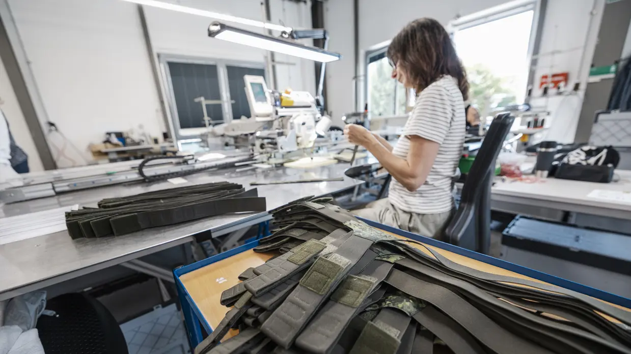 Vrouw in naaiatelier maakt militaire bretels. Een stapel bretels in groene camouflagekleur ligt op de tafel naast haar.