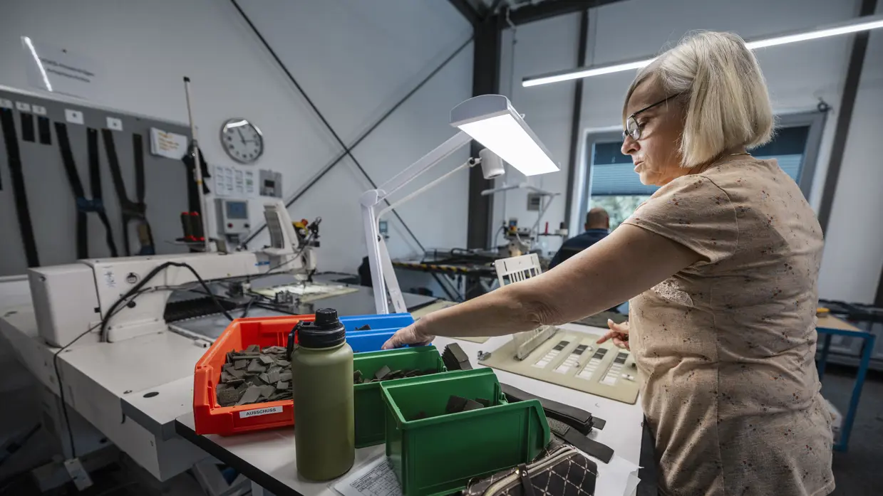 Vrouw achter tafel in atelier pakt onderdelen uit bakje voor in elkaar zetten van bretels.