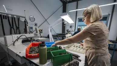 Vrouw achter tafel in atelier pakt onderdelen uit bakje voor in elkaar zetten van bretels.