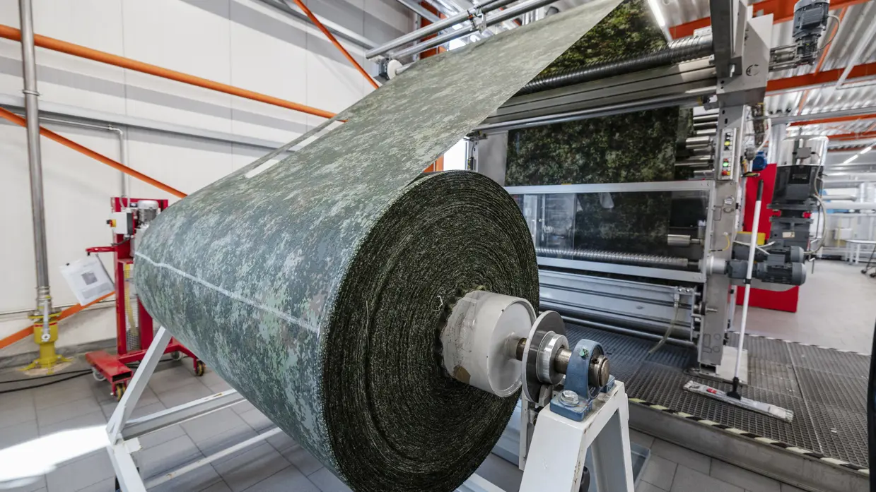 Grote rol stof in camouflagepatroon wordt door machine in productiehal geleid.
