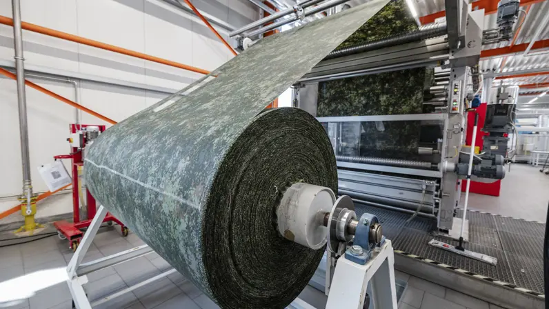Grote rol stof in camouflagepatroon wordt door machine in productiehal geleid.