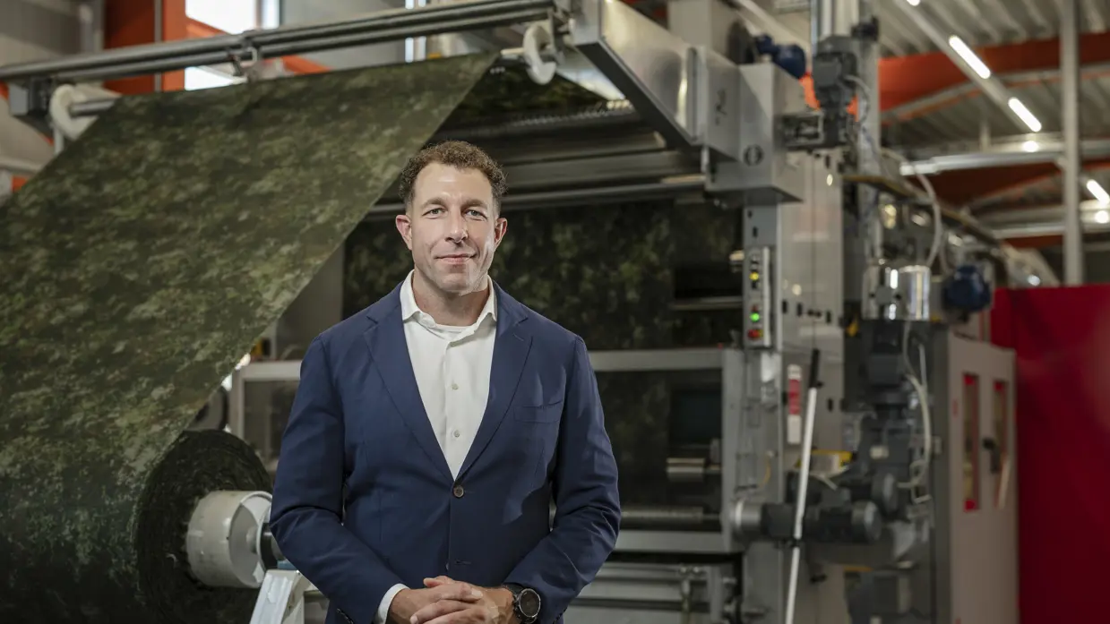 Man met blauw colbertjas poseert in fabriekshal. Op de achtergrond wordt een grote rol stof in camouflagepatroon door een machine geleid.