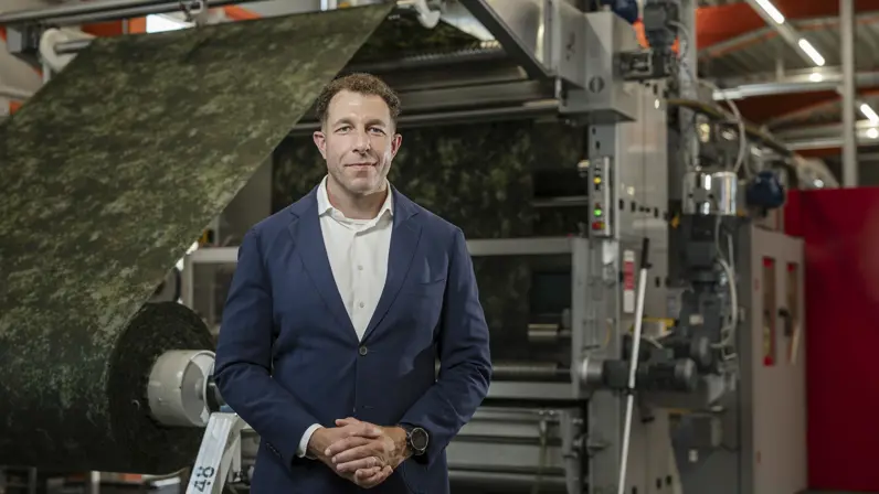 Man met blauw colbertjas poseert in fabriekshal, Op de achtergrond wordt een grote rol stof in camouflagepatroon door een machine geleid.