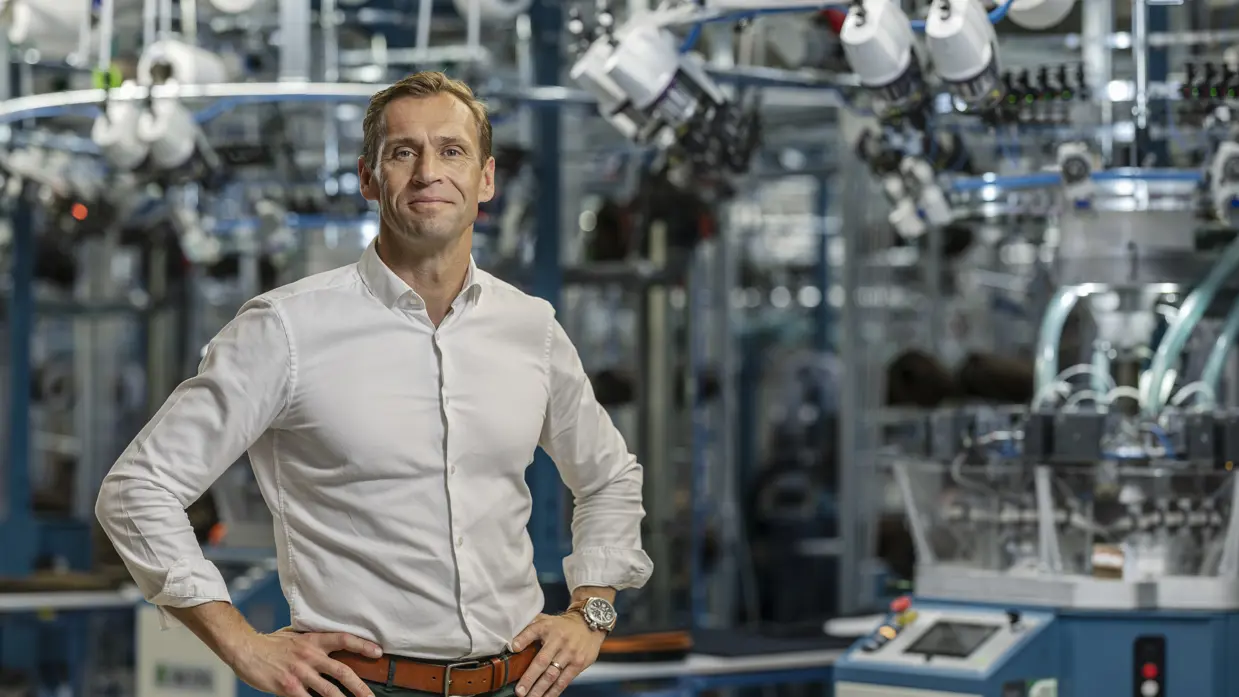 Man in witte blouse poseert met handen in de zij in fabriekshal, Op de achtergrond staan grote machines.