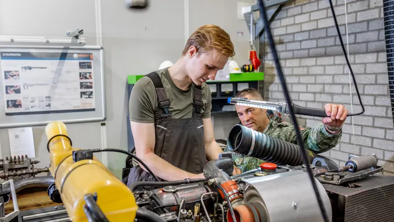 een monteur werkt aan een motor, terwijl een collega met een lamp bij schijnt. 
