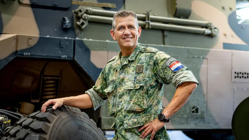 de adjudant die in camouflagekleding met een hand op een grote band leunt. 