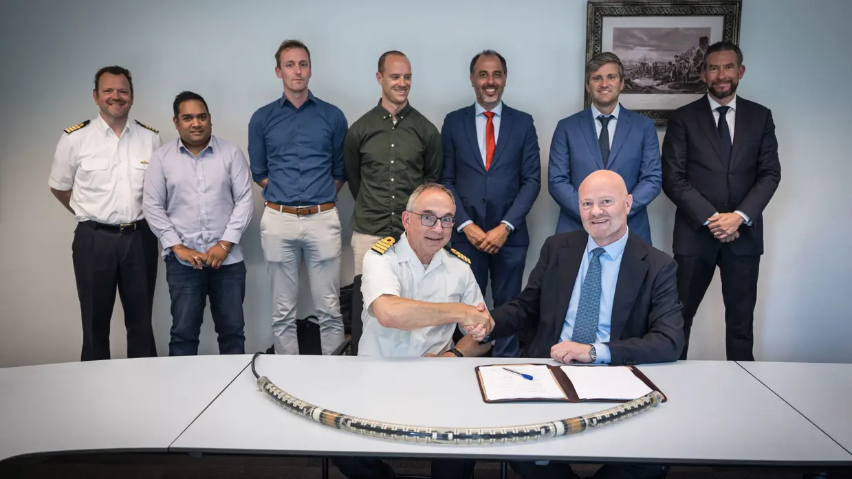 De ondertekenaars zitten achter een tafel met mensen er achter en het systeem er op.