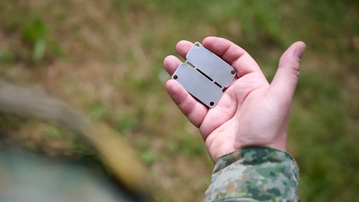 Arm van militair in groen vlekkenpak met in de hand een zilvergrijs herkenningsplaatje zonder tekst.