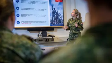 Militair in vergaderruimte staat voor groot scherm tijdens presentatie aan collega’s in diezelfde ruimte. 