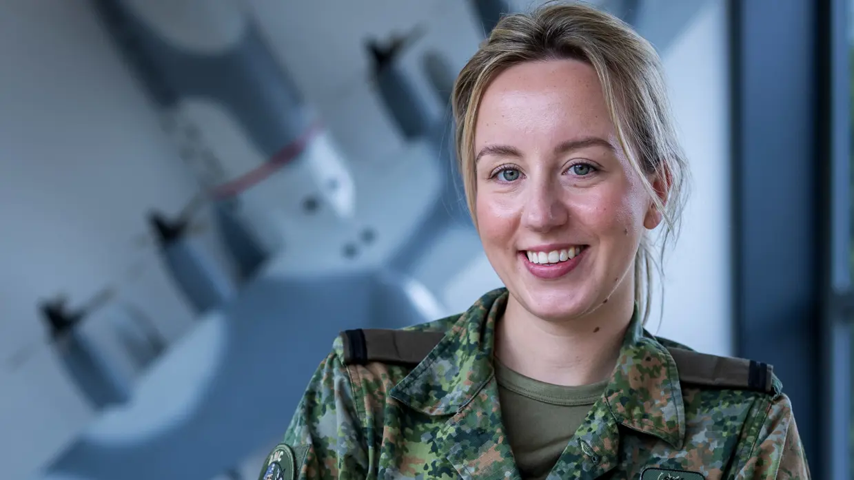 Militair kijkt lachend in de camera terwijl ze tegen reling leunt. Op de achtergrond hangt een schaalmodel van een vliegtuig aan de muur.