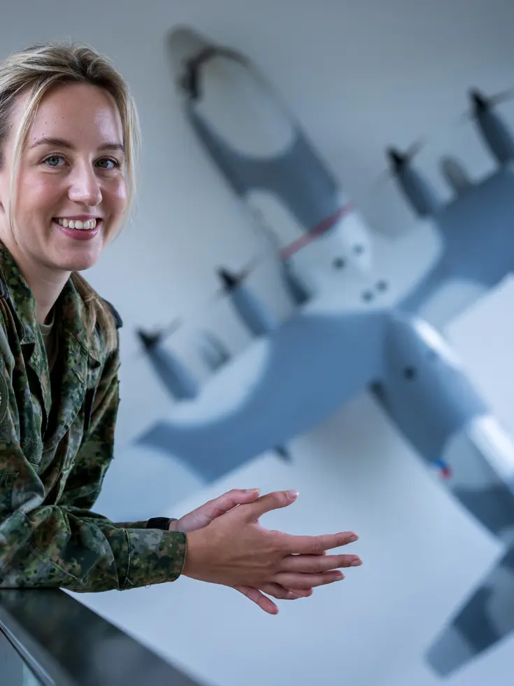 Militair kijkt lachend in de camera terwijl ze over reling leunt. Op de achtergrond hangt een schaalmodel van een C-130 Hercules aan de muur.