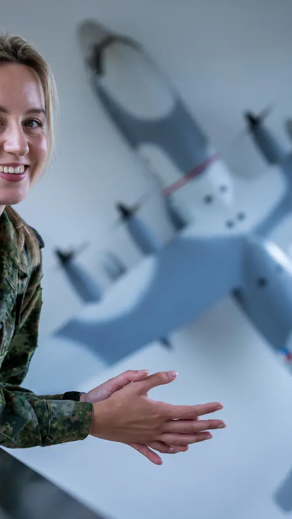Militair kijkt lachend in de camera terwijl ze over reling leunt. Op de achtergrond hangt een schaalmodel van een C-130 Hercules aan de muur.