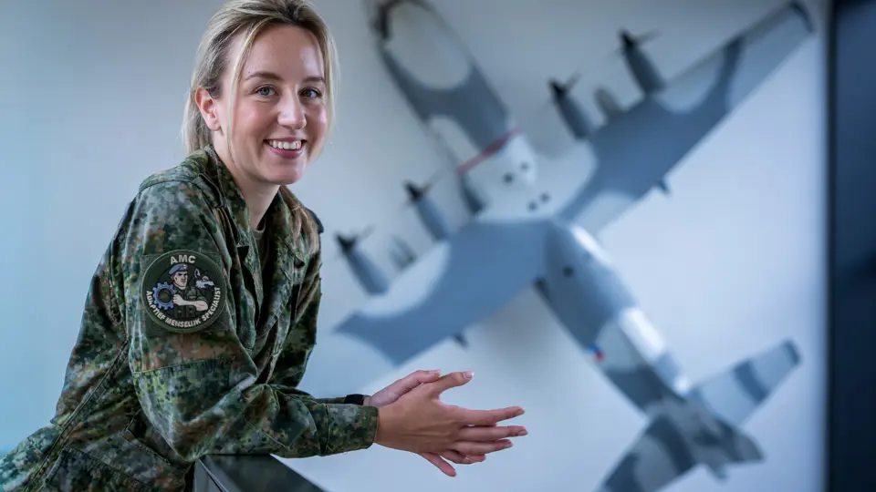 Militair kijkt lachend in de camera terwijl ze over reling leunt. Op de achtergrond hangt een schaalmodel van een C-130 Hercules aan de muur.