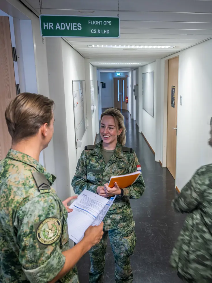 Militair in kantoorgang in gesprek met collega-militair terwijl ze paperassen vasthouden.