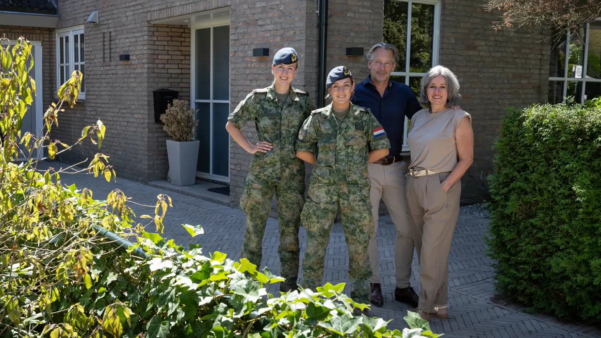 Vader en moeder in burgerkleding staan naast 2 dochters in militair uniform en poseren allen glimlachend voor een bakstenen huis met garage en scooter.