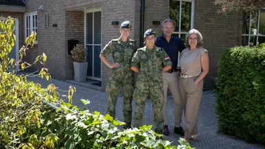 Vader en moeder in burgerkleding staan naast 2 dochters in militair uniform en poseren allen glimlachend voor een bakstenen huis met garage en scooter.