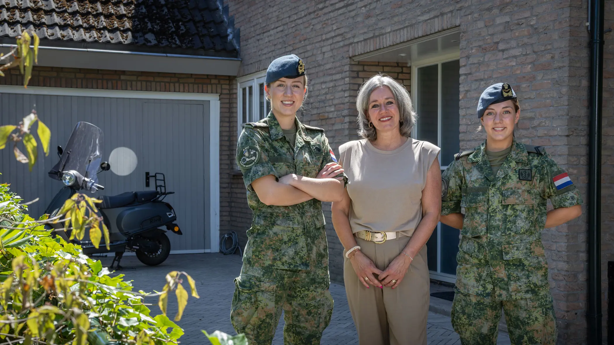 Moeder in burgerkleding staat tussen twee dochters in militair uniform en poseren allen glimlachend voor een bakstenen huis met garage en scooter.