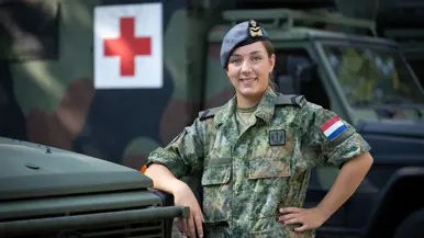 Militair in uniform leunt op motorkap ambulance. Op de achtergrond een groot rood kruis op de zijkant van een andere ambulance.