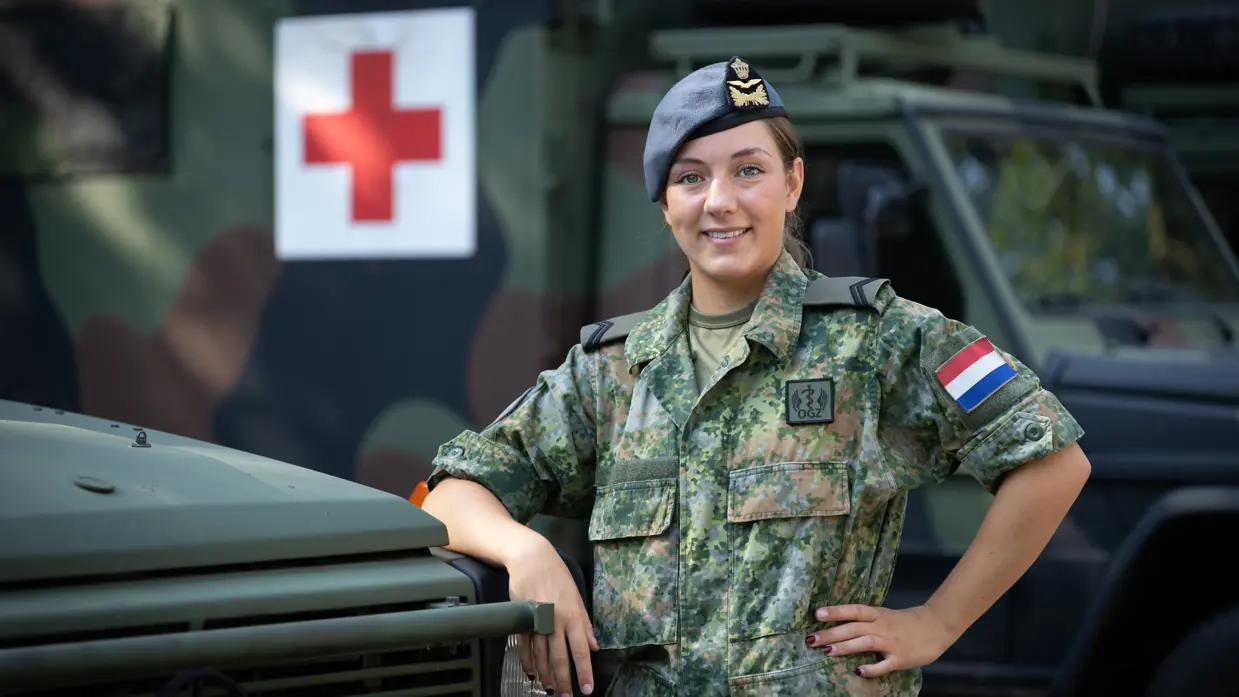 Militair in uniform leunt op motorkap ambulance. Op de achtergrond een groot rood kruis op de zijkant van een andere ambulance.