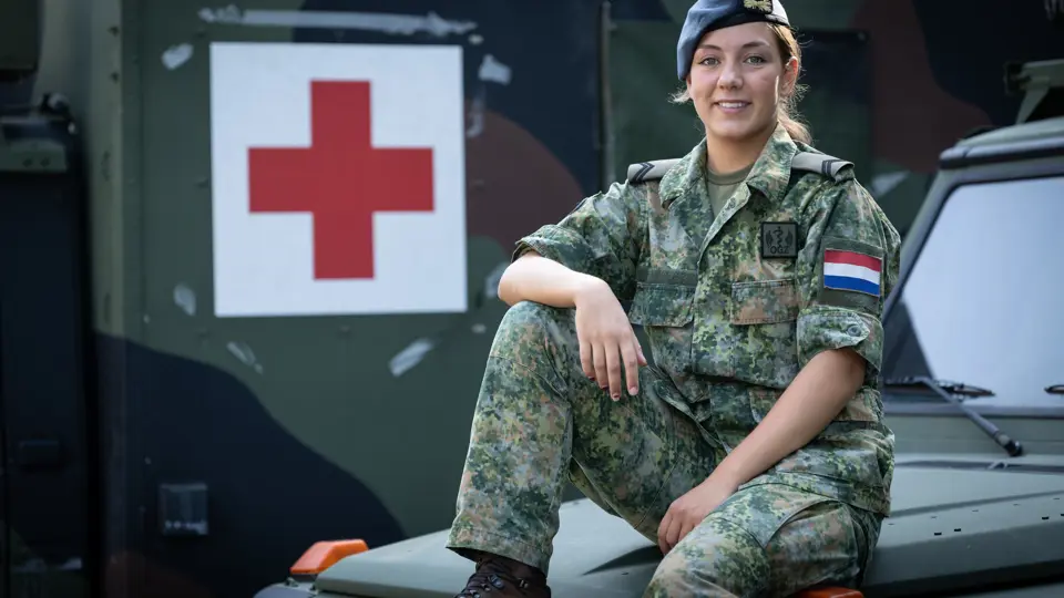 Militair in uniform zit op motorkap ambulance. Op de achtergrond een groot rood kruis op de zijkant van een andere ambulance.