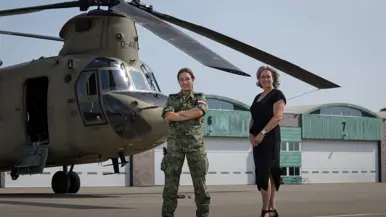 Militair en burgermedewerker staan voor Chinook. In de lucht in de achtergrond vliegt een NH90.