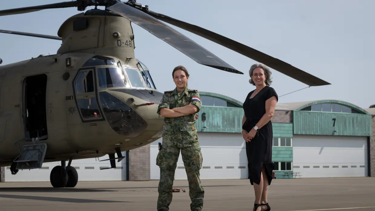Militair en burgermedewerker staan voor Chinook. In de lucht in de achtergrond vliegt een NH90.