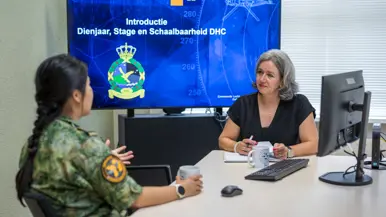 Vrouw in burgerkleding aan bureau in gesprek met militair in uniform.