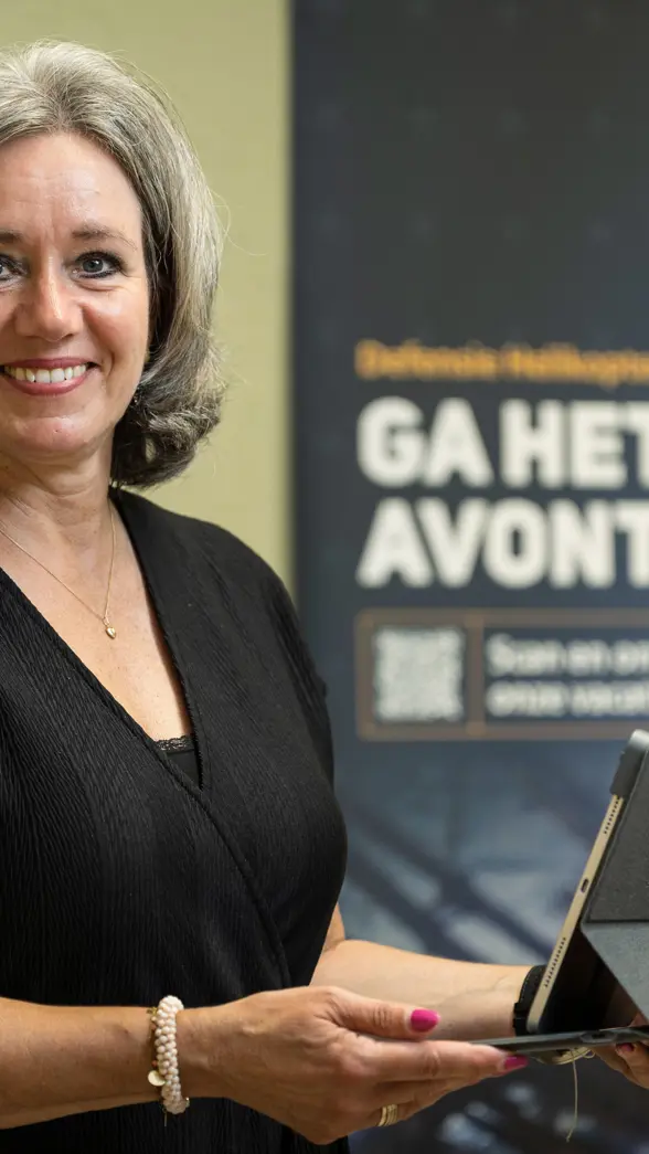 Vrouw staat met tablet in de hand en kijkt lachend in de camera.