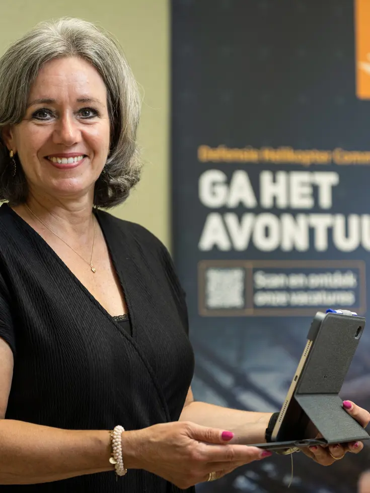 Vrouw staat met tablet in de hand en kijkt lachend in de camera.