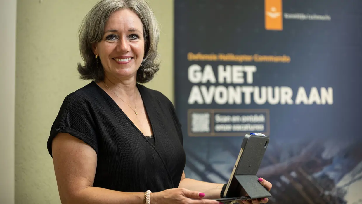 Vrouw staat met tablet in de hand en kijkt lachend in de camera.