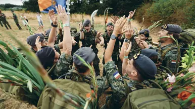 Juichende militairen in een kringetje op een veld. Ze dragen een gevechtstenue met rugzak waaraan gladiolen vastzitten.