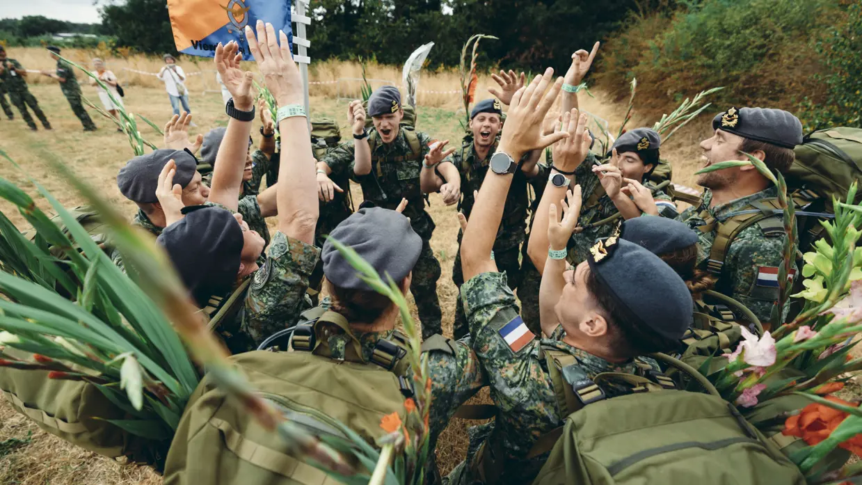 Juichende militairen in een kringetje op een veld. Ze dragen een gevechtstenue met rugzak waaraan gladiolen vastzitten.