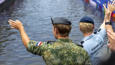 Twee militairen voorop de boot op de rug bekeken zwaaien naar de kades links en rechts waar een gekleurde mensenmenigte staat.