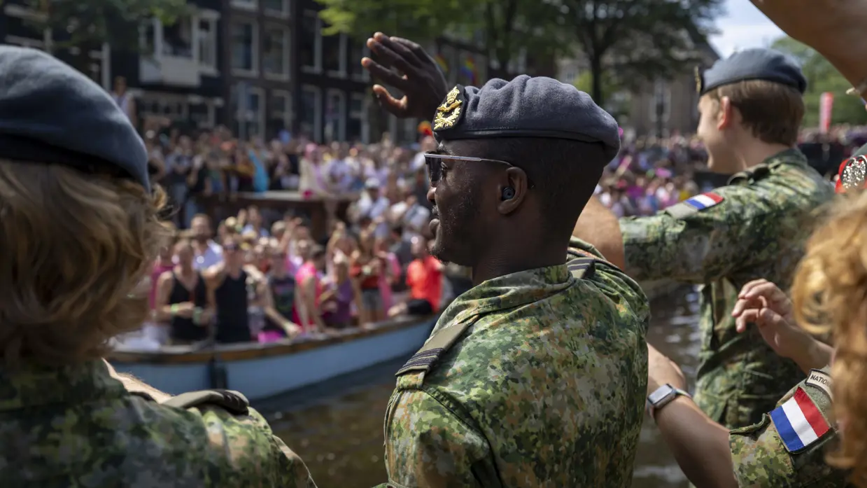 Drie luchtmachtmilitairen vanaf de zij- en achterkant bekeken zwaaien vanaf een boot naar een gekleurde mensenmenigte op de kade.