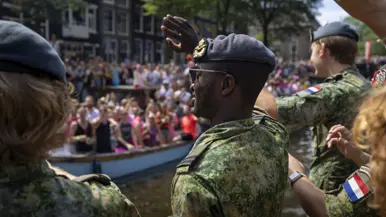Drie luchtmachtmilitairen vanaf de zij- en achterkant bekeken zwaaien vanaf een boot naar een gekleurde mensenmenigte op de kade.