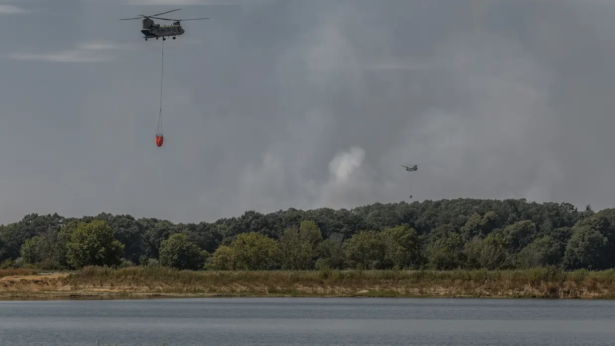 Een Chinook met Bambi Bucket boven water met op de achtergrond natuurgebied waarboven een tweede Chinook vliegt. Er is lichte rook boven de begroeiing te zien.