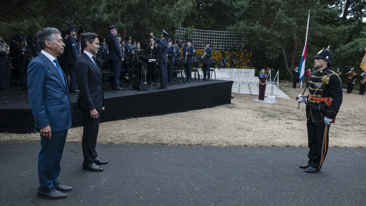 Jetten staat samen met een man in burgerkleding op afstand tegenover een militair in ceremonieel tenue. Op de achtergrond speelt het Orkest Koninklijke Luchtmacht naast het Indisch Monument.