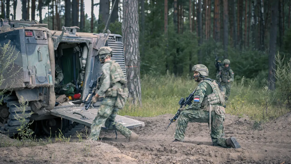 Drie soldaten in camouflagekleding bij een pantservoertuig in een bos, één loopt, één knielt met een wapen.