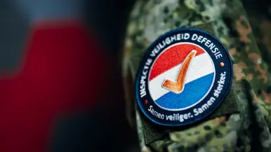 Een badge van de Inspectie Veiligheid Defensie met de kleuren van de Nederlandse vlag met daarop een oranje vinkje. De slogan luidt: Samen veiliger. Samen sterker.