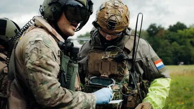 2 militairen met helm, bril en geluidsapparatuur overleggen op een weide met elkaar.
