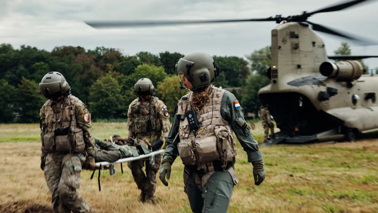3 militairen met helm, bril, vizier en bepakking tillen een brancard met een militair erop en lopen op een weide weg van een helikopter.