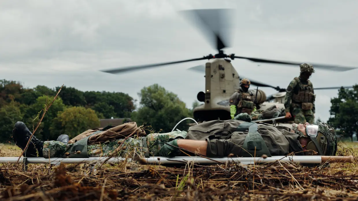 Een gewonde militair ligt op een weide op een brancard, terwijl achter hem een andere militair per brancard wordt afgevoerd door 2 militairen. Daarachter staat een helikopter.