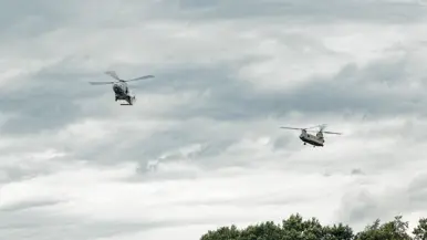 2 helikopters vliegen achter elkaar ver boven de bomen.