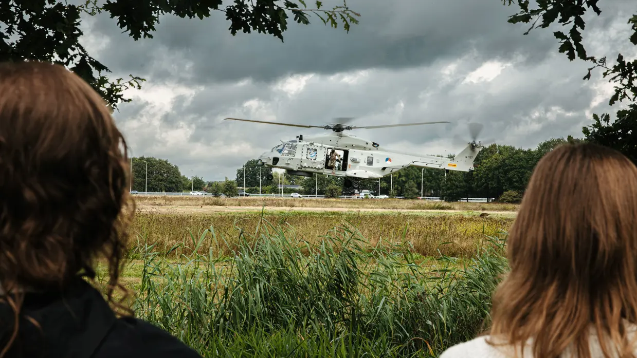 Een helikopter met openstaande deuren en een militair bij de uitgang landt op een weide met toeschouwers op afstand.