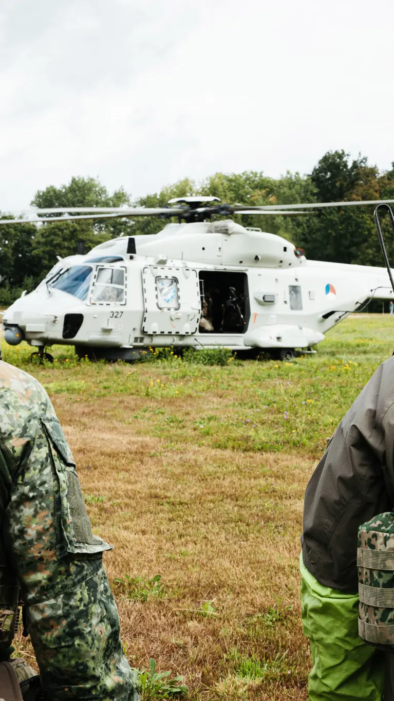 2 bepakte militairen met helmen op staan op een weide tegenover een helikopter met openstaande deuren.