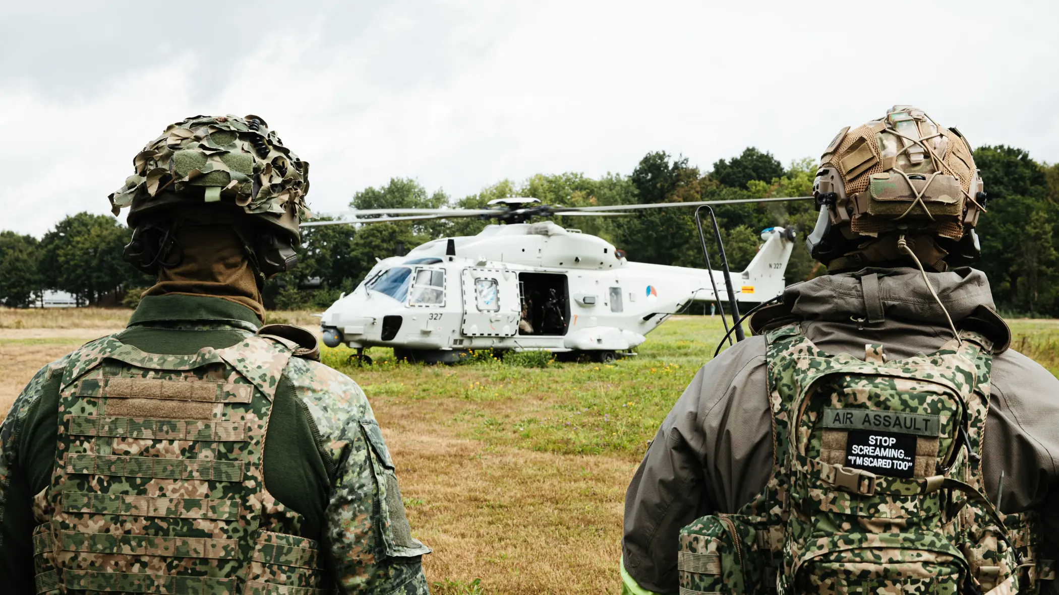 2 bepakte militairen met helmen op staan op een weide tegenover een helikopter met openstaande deuren.